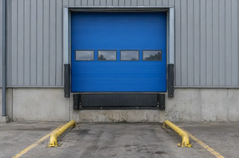 Blue insulated sectional loading bay door with vision panels above a dock leveller and rubber bumpers
